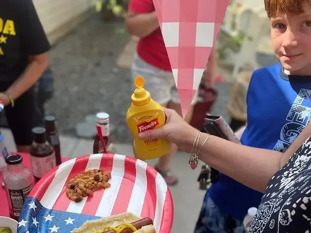 A child holding a mustard bottle over a flag-print plate at a cookout